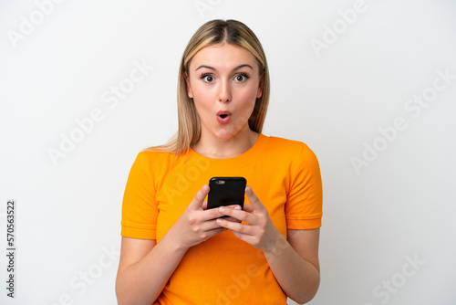 Young caucasian woman isolated on white background looking at the camera while using the mobile with surprised expression photo