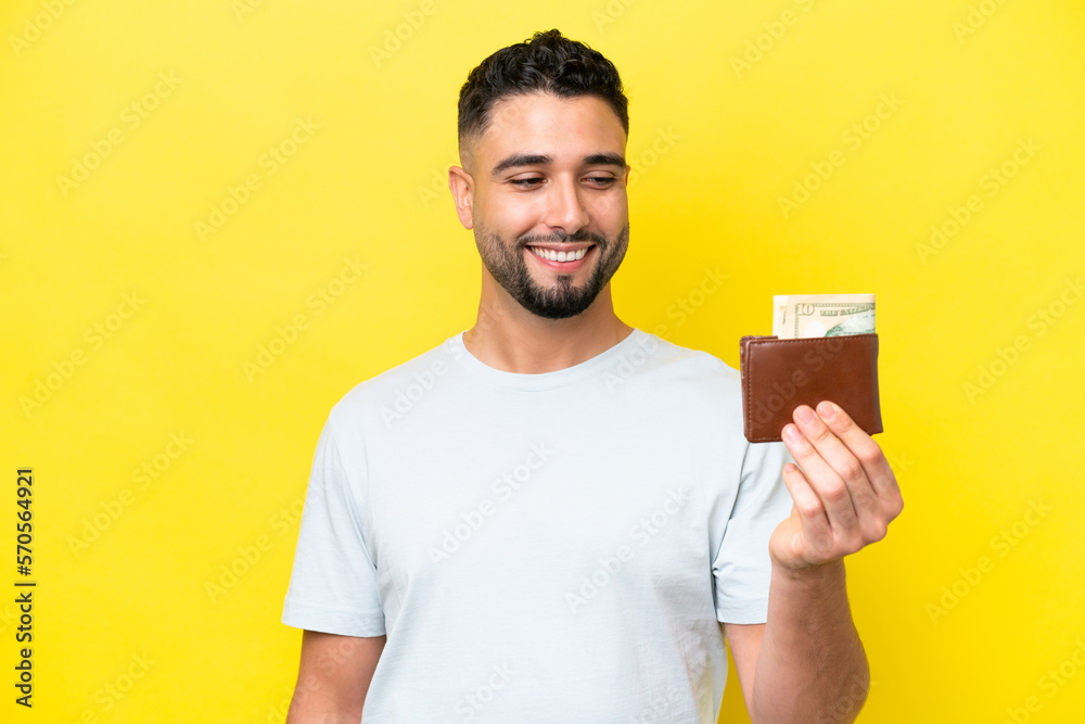 Young Arab man holding a wallet isolated on yellow background with happy expression Young Arab man holding a wallet isolated on yellow background with happy expression