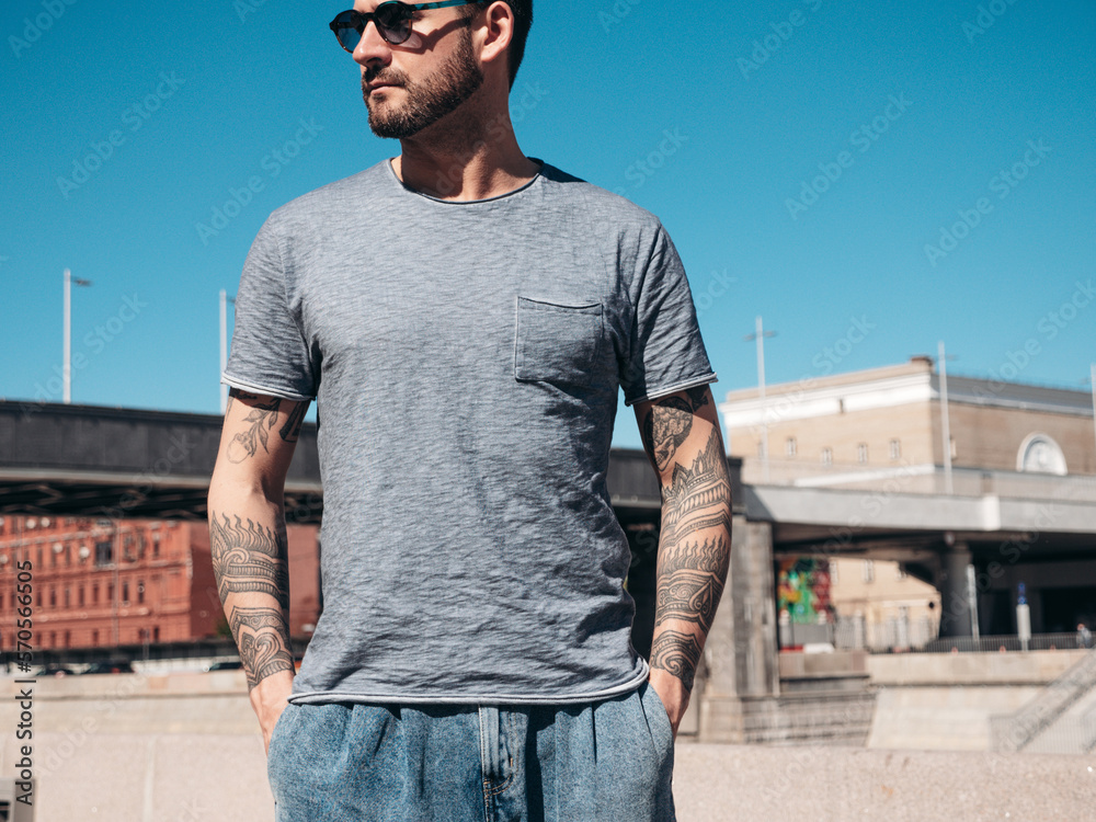 Portrait of handsome confident stylish hipster model. Sexy man dressed in jeans and grey t-shirt. Fashion male posing in the street. In Europe city. Outdoors at sunny day. With tattoos Portrait of handsome confident stylish hipster model. Sexy man dressed in jeans and grey t-shirt. Fashion male posing in the street. In Europe city. Outdoors at sunny day. With tattoos