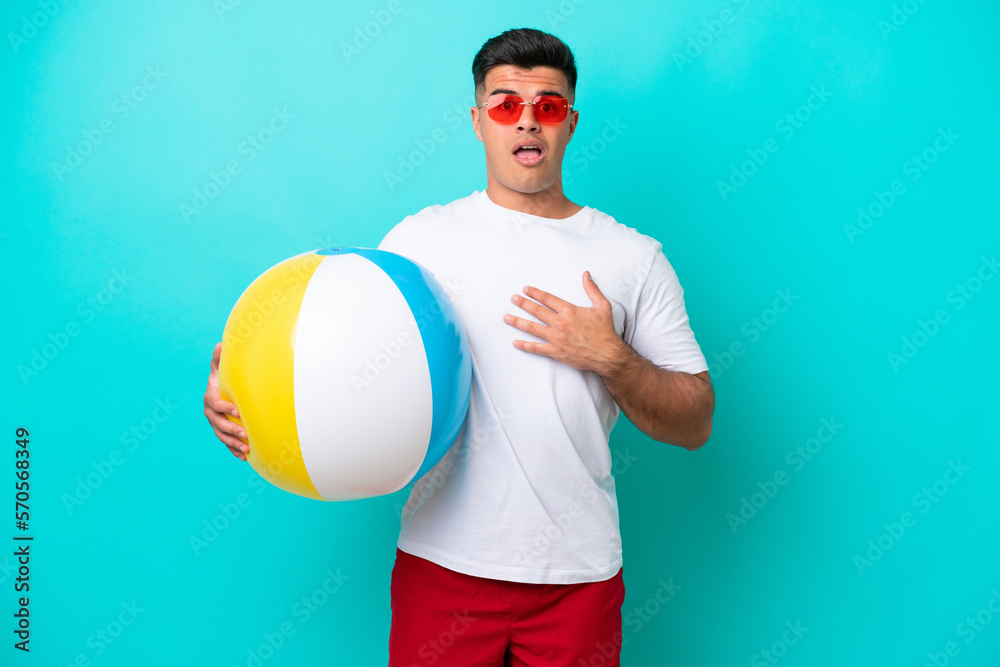 Young caucasian man holding a beach ball isolated on blue background surprised and shocked while looking right