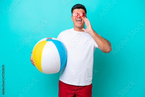 Young caucasian man holding a beach ball isolated on blue background shouting with mouth wide open