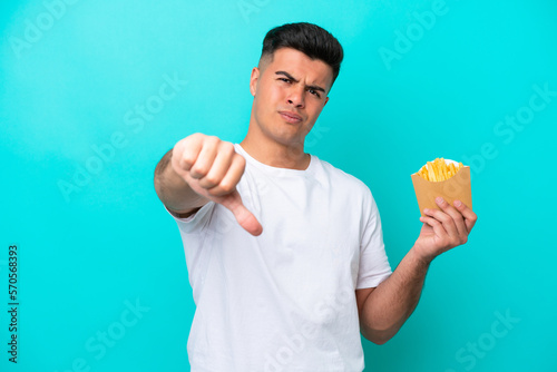 Young caucasian man catching french fries isolated on blue background showing thumb down with negative expression © luismolinero