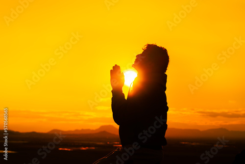 Silhouette of a person with joined hands, looking at the sky and praying. The sun is between his hands and his face, crossing his body with the rays of the sun. Religion and prayer concept.