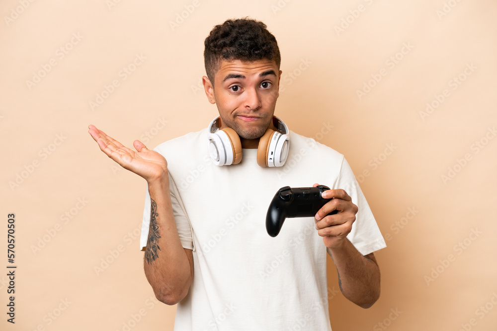 Young Brazilian man playing with a video game controller isolated on beige background having doubts while raising hands Young Brazilian man playing with a video game controller isolated on beige background having doubts while raising hands