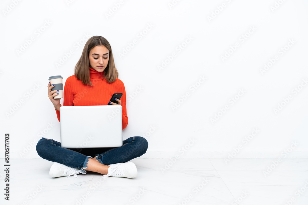 Young caucasian woman sitting on the floor with a laptop holding coffee to take away and a mobile Young caucasian woman sitting on the floor with a laptop holding coffee to take away and a mobile