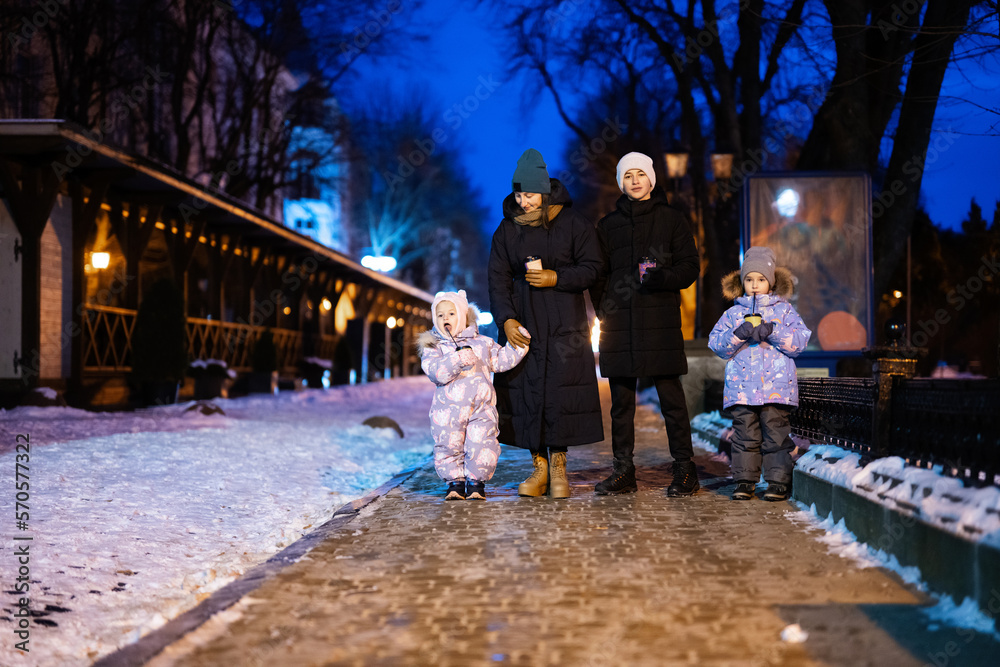 Family walk through the night city in the winter. A woman and three children in evening drink hot drink in cups.