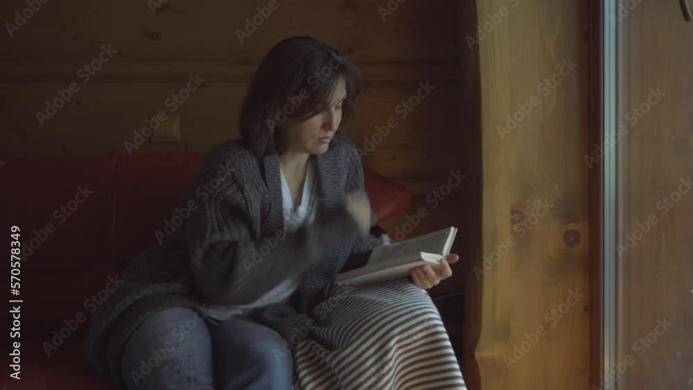 Woman sitting on cozy comfortable sofa near window and reading book. Spending time at home, coziness 4K video concept.