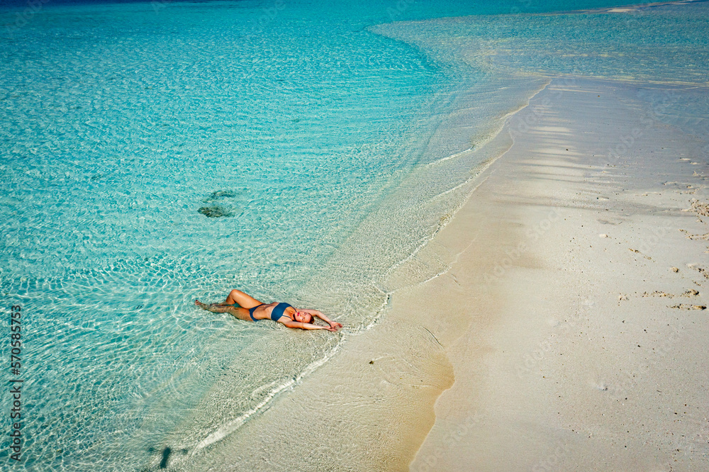 A young woman in a blue bikini lies on her back on the sand near the waves of the blue sea. View from above. The beach in the Maldives, clear clean water and yellow sand