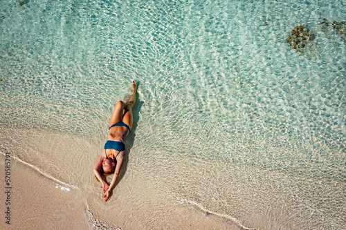 A young woman in a blue bikini lies on her back on the sand near the waves of the blue sea. View from above. The beach in the Maldives, clear clean water and yellow sand