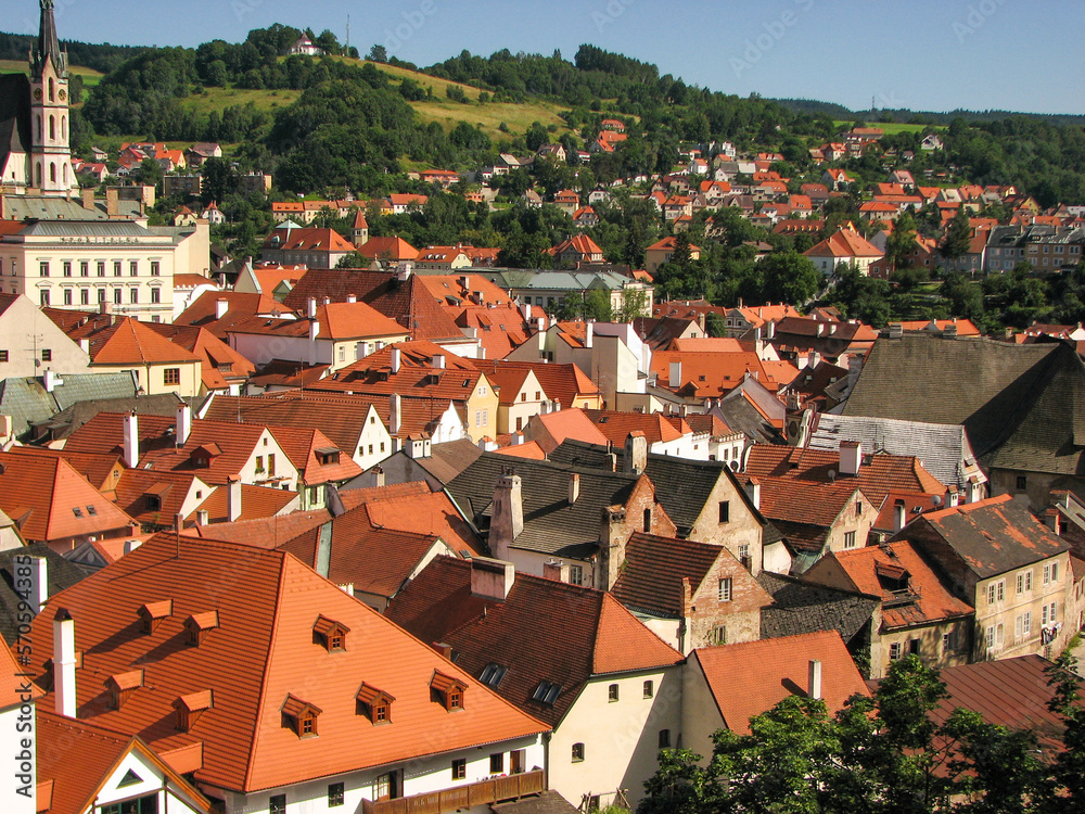 An aerial view of Cesky Krumlov’s old town, including several siena-coloured rooftops and a church tower on a sunny day with a blue sky.  Image has copy space.
