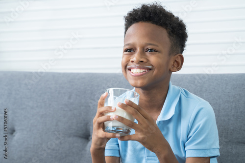 Portrait of cheerful African Nigerian boy smiling with milk moustache, holding and drinking a glass of milk, sitting on sofa at home. Healthy food and drink for children concept