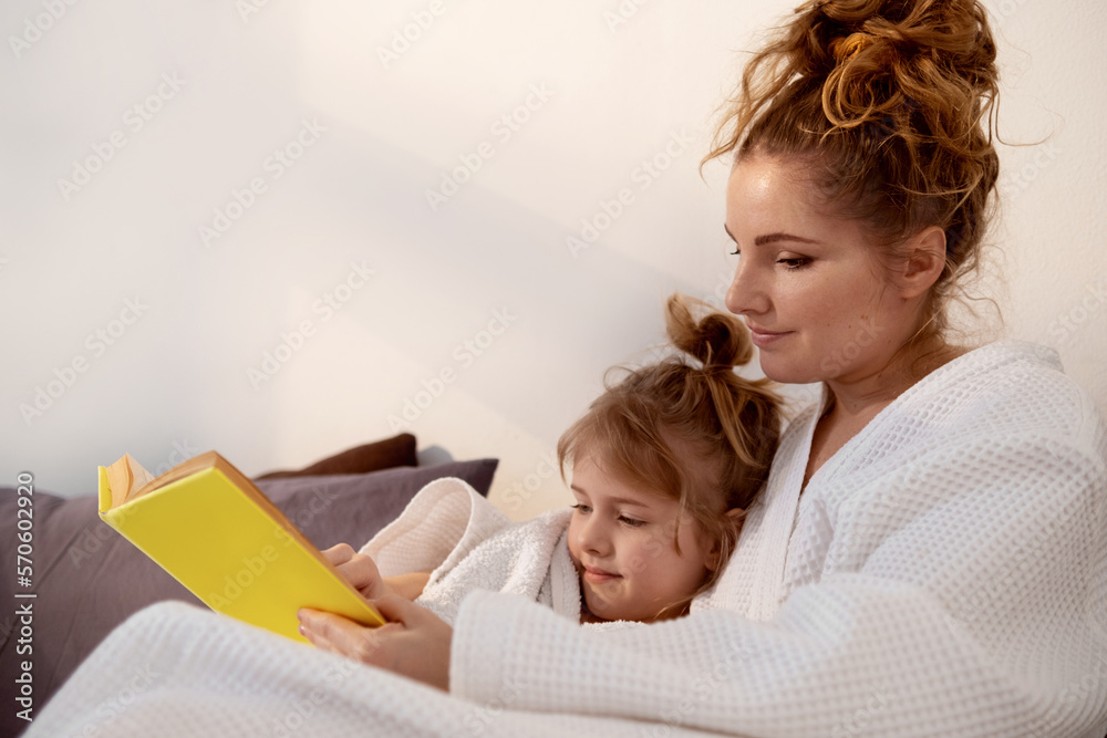 Mother reading a book to her little daughter in a cozy bed