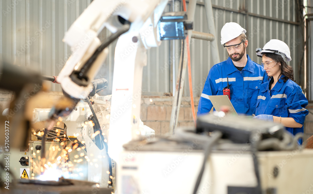 Industrial engineer using laptop computer to control robotic welder arm operation in modern automation factory. Woman technician use robot machine to do automated steel welding full of fire sparks Industrial engineer using laptop computer to control robotic welder arm operation in modern automation factory. Woman technician use robot machine to do automated steel welding full of fire sparks