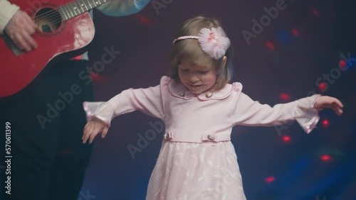Little girl on stage in pink vintage dress, she dances, her father plays acoustic guitar. Color music is shining and smoke is billowing. Father Day. Performance on stage. Hobbies and creativity