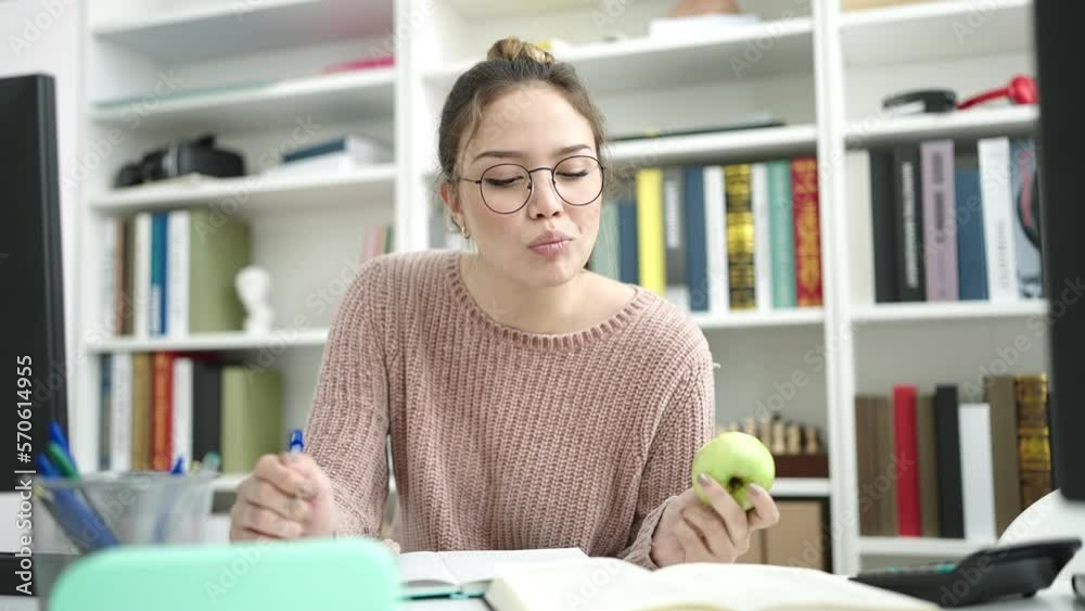 Young beautiful hispanic woman student writing on notebook eating apple at library university