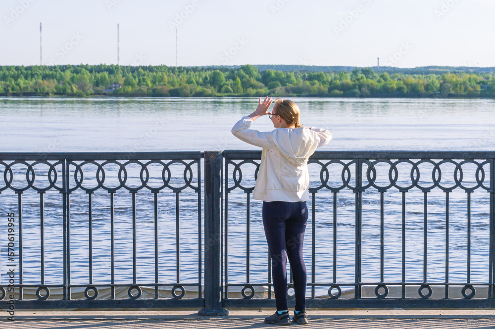 A young girl stands on the embankment and looks towards the river. A girl's walk by the river on a summer day. A blonde girl in a white windbreaker with her arms thrown back looks into the distance.
