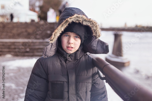 Portrait of a young boy in winter clothes in winter in the city, the boy is sad because he is cold, a strong wind is blowing near the river. The boy needs warmth and protection
