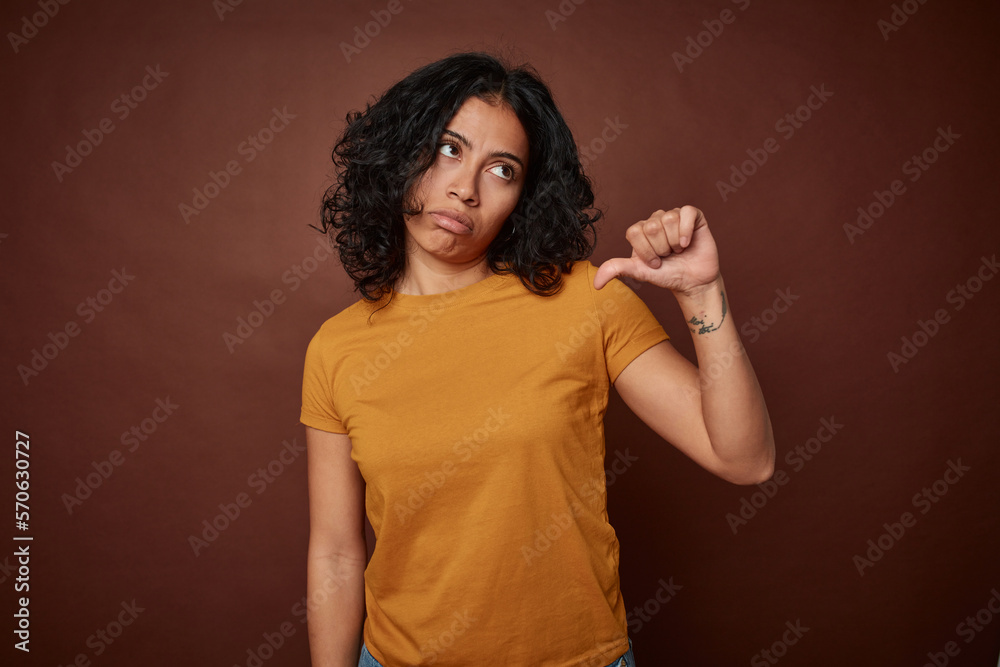 Young colombian curly hair woman isolated on brown background showing a dislike gesture, thumbs down. Disagreement concept. Young colombian curly hair woman isolated on brown background showing a dislike gesture, thumbs down. Disagreement concept.