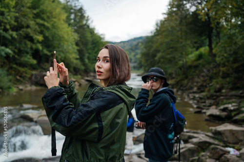 A young couple of tourists are standing in the mountains near the river against the background of a beautiful landscape, taking a photo on a smartphone camera and looking into the camera