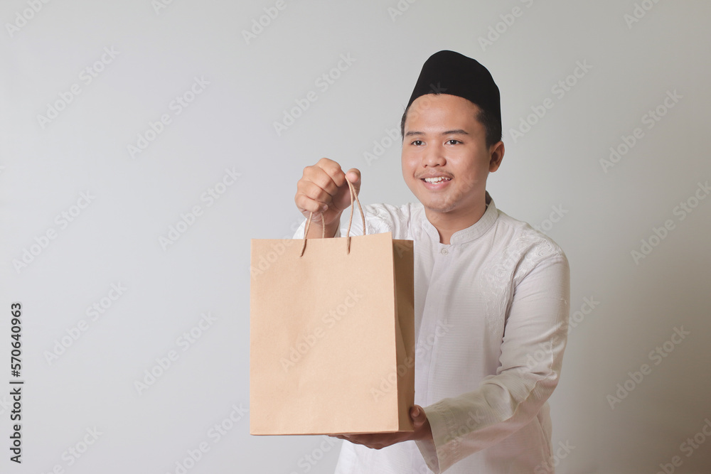 Portrait of Asian muslim man in white koko shirt with skullcap giving craft paper shopping bag with gift inside. Isolated image on gray background Portrait of Asian muslim man in white koko shirt with skullcap giving craft paper shopping bag with gift inside. Isolated image on gray background