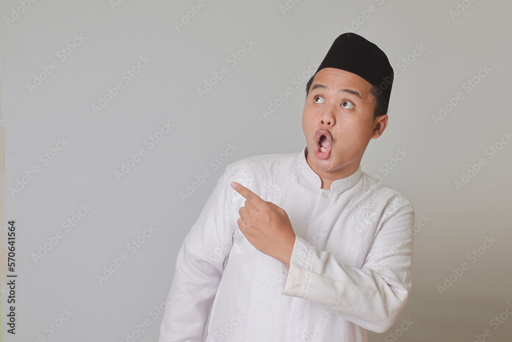 Portrait of Asian muslim man in white koko shirt with skullcap poiting to the side and showing wow face expression. Isolated image on gray background Portrait of Asian muslim man in white koko shirt with skullcap poiting to the side and showing wow face expression. Isolated image on gray background