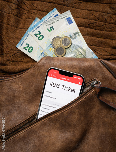 Aus einem geöffneten Reißverschluss einer braunen Handtasche schaut ein Smartphone heraus, auf dem gerade eine Fahrkarten-App mit einem 49€-Ticket darauf geöffnet ist. Im Hintergrund braun Strick