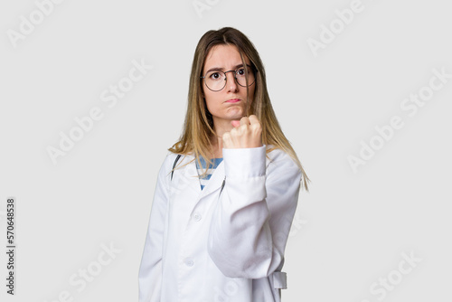 Compassionate female physician with a stethoscope around her neck, ready to diagnose and care for her patients in her signature white coat showing fist to camera, aggressive facial expression.