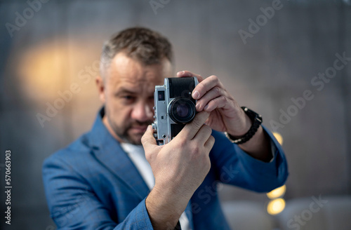 A bearded man in a suit holds a vintage film camera. Shot on a gray background.