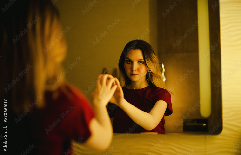 Portrait of a beautiful blonde woman with an expressive look in a burgundy T-shirt, who touches her reflection in the mirror, standing in the semi-darkness in the bathroom. Portrait of a beautiful blonde woman with an expressive look in a burgundy T-shirt, who touches her reflection in the mirror, standing in the semi-darkness in the bathroom.