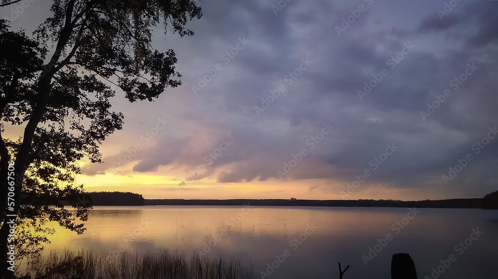 Sunset sky over lake landscape, nature background with heavy clouds and tree silhouette