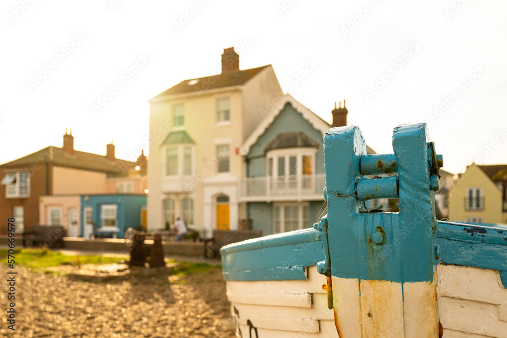Shallow focus of an old wooden fishing boat seen on a shingle shoreline. Coastal properties can be seen in the summer light. Shallow focus of an old wooden fishing boat seen on a shingle shoreline. Coastal properties can be seen in the summer light.