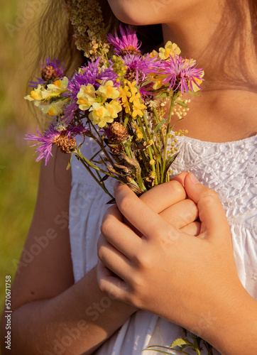 Teen girl wearing white dress holding wild flowers bouquet on a grassy sunny summer meadow field