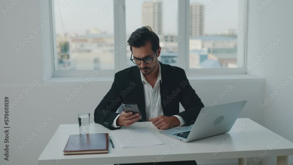 Man business looking in phone and smiling, news, working in office typing at laptop, work day and paperwork, technology and online. Man business looking in phone and smiling, news, working in office typing at laptop, work day and paperwork, technology and online.