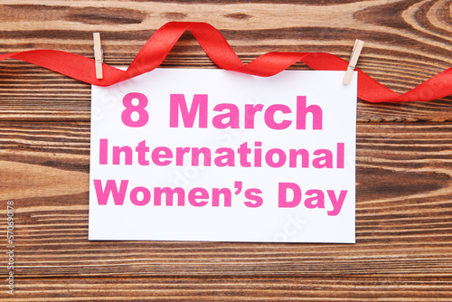 Card with text 8 March International Women's Day hanging on red ribbon, on brown wooden background