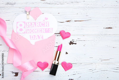 Card in shape of heart with text 8 March Women's Day in envelope. Pink ribbon and hearts, and lipstick on white wooden background