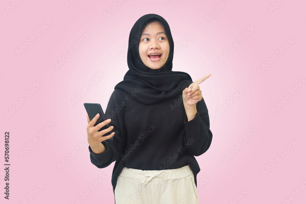 Portrait of excited Asian hijab woman in black outfit showing product, pointing away with her finger while using mobile phone. Advertising concept. Isolated image on pink background Portrait of excited Asian hijab woman in black outfit showing product, pointing away with her finger while using mobile phone. Advertising concept. Isolated image on pink background