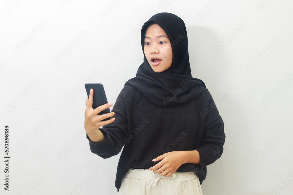 Portrait of shocked Asian hijab woman in black outfit looking at her mobile phone. Wow face expression. Advertising concept. Isolated image on white background Portrait of shocked Asian hijab woman in black outfit looking at her mobile phone. Wow face expression. Advertising concept. Isolated image on white background