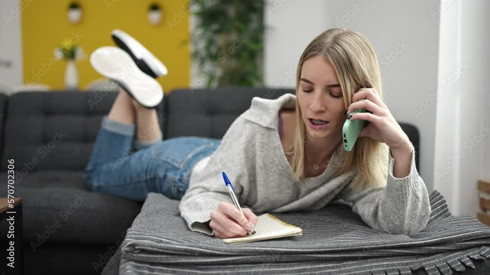 Young blonde woman writing on notebook lying on sofa speaking on the phone at home Young blonde woman writing on notebook lying on sofa speaking on the phone at home