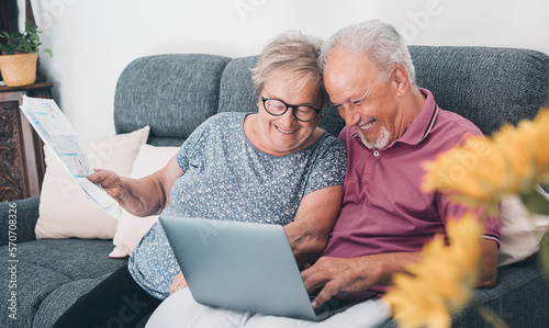 Aged couple at home paying bills on line with laptop and laughing a lot having fun together. Happiness and elderly lifestyle. Man and woman old senior sitting on sofa with computer online connection photo