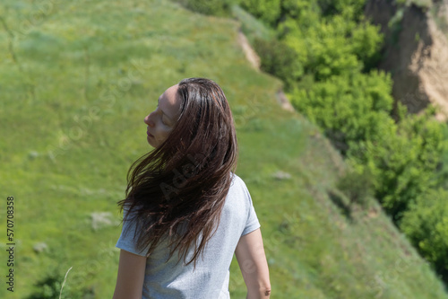 Portrait woman with long brown hair. Person posingin standing on ravine covered with greenery. Confident young adult female makes head movement and wind blowing hair. Enjoyment of life in 35-45 years.