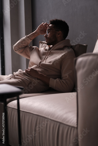 Portrait of a cheerful young african american man 20s wearing casual hoody and glasses sitting on sofa against black background. copy space isolated