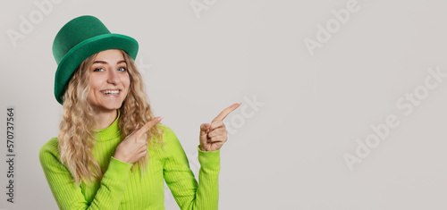 St. Patrick's Day leprechaun model girl in green hat  isolated on white background pointing to the right and smiling, having fun. Patrick Day pub party, celebrating.