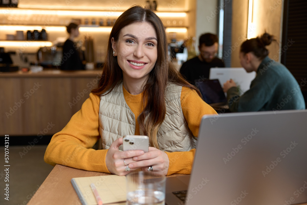 Horizontal shot of beautiful caucasian female smiling dressed in casual yellow sweater and beige vest using a smartphone working on laptop looking at camera posing while sitting in a cafe indoors. Horizontal shot of beautiful caucasian female smiling dressed in casual yellow sweater and beige vest using a smartphone working on laptop looking at camera posing while sitting in a cafe indoors.