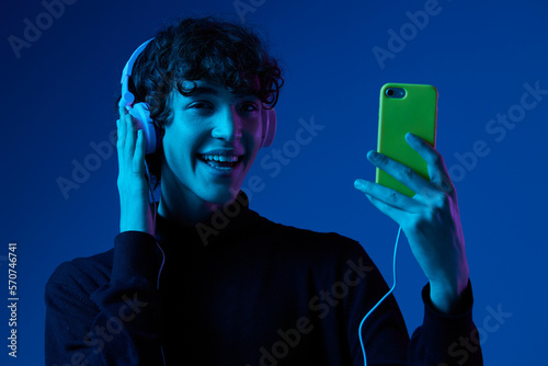 Man smile with phone in hand looking at the smartphone screen in headphones listening to music, portrait dark blue background, neon light, style and trends, mixed light, men's fashion, copy space © SHOTPRIME STUDIO