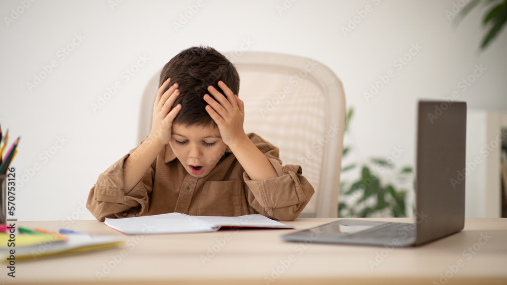 Shocked sad european small kid with open mouth hold head with hands, has difficulties with study in living room Shocked sad european small kid with open mouth hold head with hands, has difficulties with study in living room