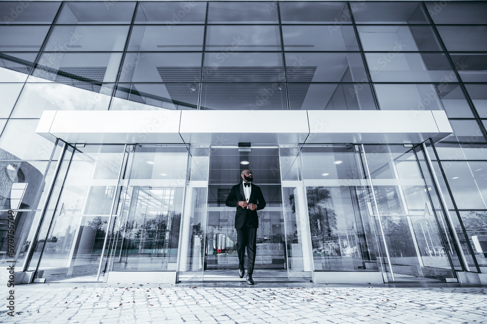 A wide-angle shot of a black man looking dapper in a full-grown beard exudes elegance in his black tuxedo, bow tie, and varnished leather shoes as he leaves a modern glass building