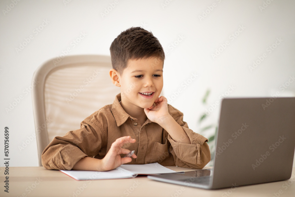 Elementary education remotely. Smiling caucasian little child sit at table, make notes, watch lesson on computer Elementary education remotely. Smiling caucasian little child sit at table, make notes, watch lesson on computer