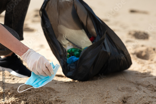 Problem of ocean polution after coronavirus pandemic. Female volunteer picking up trash and medical face masks on beach © Prostock-studio