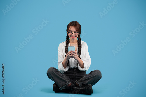Smiling caucasian teen girl with pigtails student sit on floor, chatting on phone, isolated on blue background