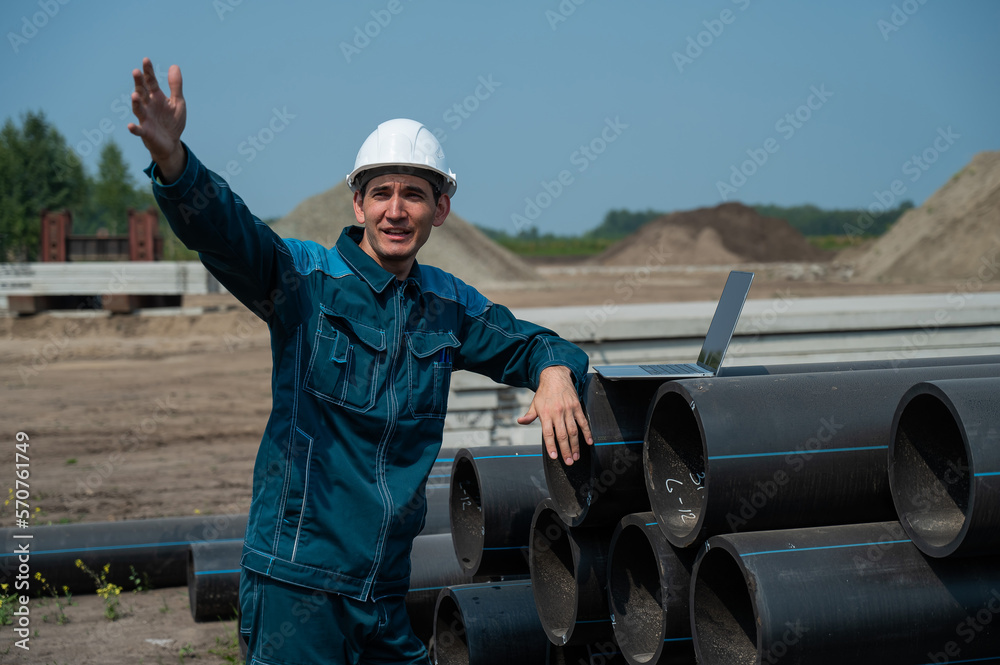 Caucasian male builder in a hard hat stands near the pipes and uses a laptop at a construction site. Caucasian male builder in a hard hat stands near the pipes and uses a laptop at a construction site.